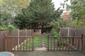 A wooden deck with a gate leading to a backyard with a large tree, a wooden fence, and a small garden area. - Spacious 4BR Townhouse for 9 near Tufnell Park & Holloway (London)