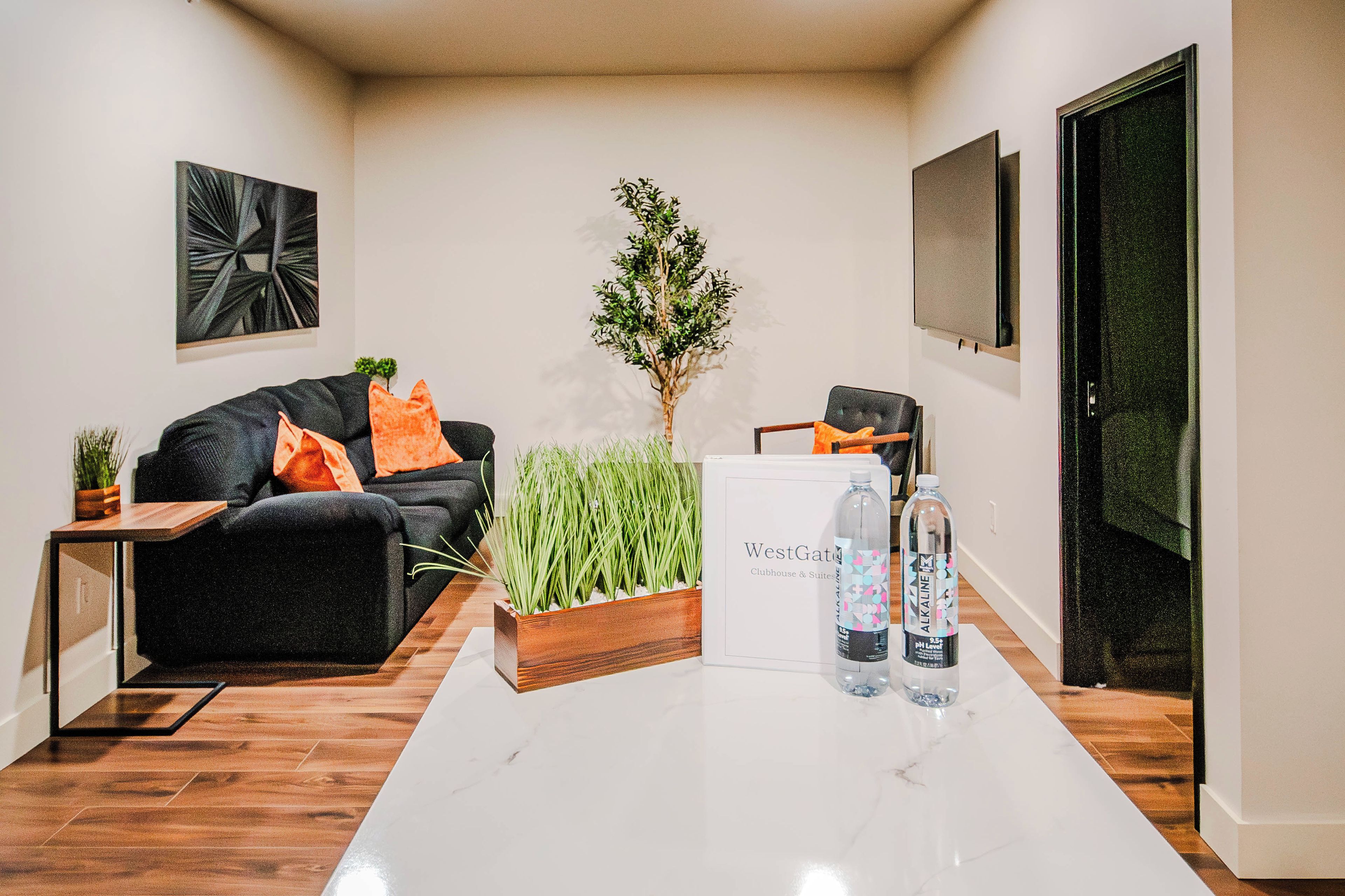 A modern living room with a black sofa, orange cushions, a wooden side table, a potted plant, and a sign for WestGate Clubhouse & Suites.