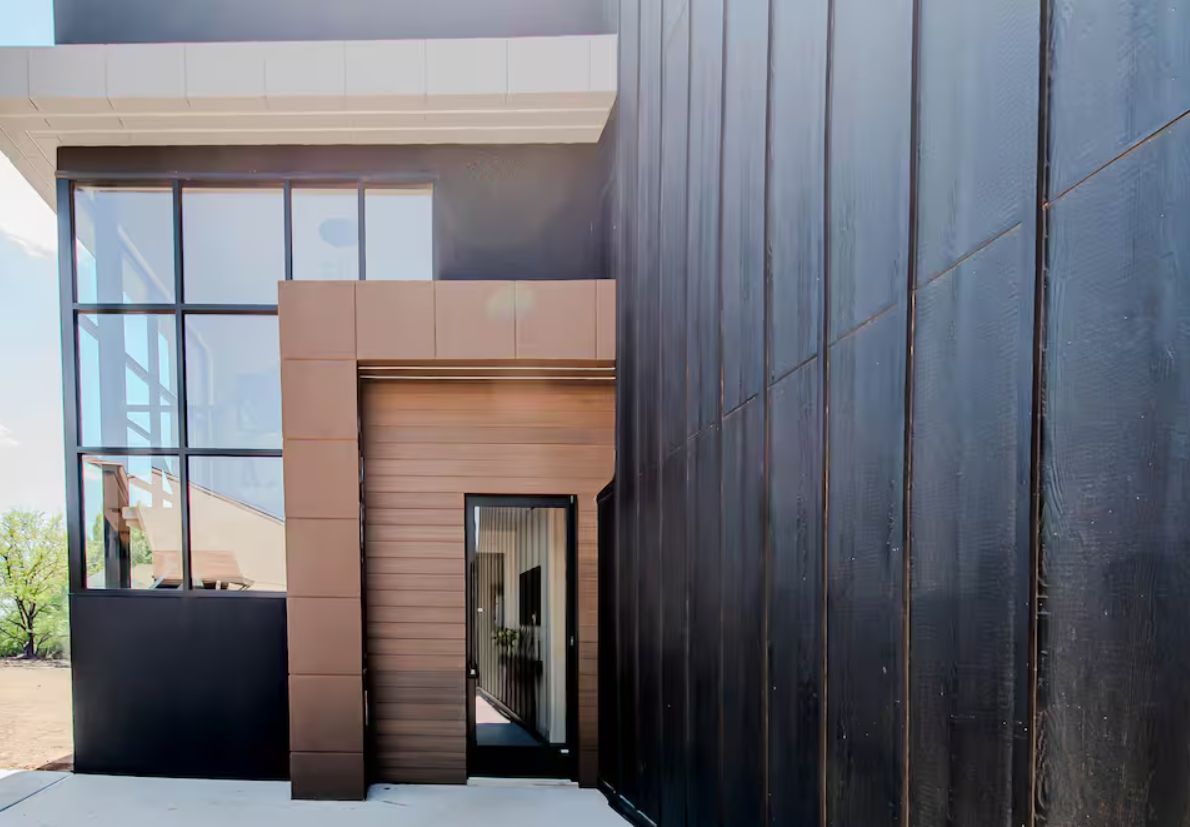 A modern building exterior with large windows, a dark facade, and a visible door leading to an interior space.