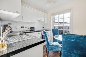 A modern kitchen with white cabinets, a black countertop, and a window with a view of a building. - Cosy 1 bed Flat in Greenwich With Private Garden (London)