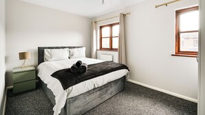 A bedroom with a bed, a nightstand, a lamp, a window with curtains, and a radiator. - Apartment Sleeps 5 With Garden (London)