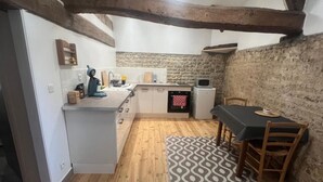 A compact kitchen with wooden beams, a stone wall, and a dining area with a patterned rug. - Apartment 'Gîte 1' with Shared Terrace, Shared Garden and Wi-Fi (Carentan-les-Marais)