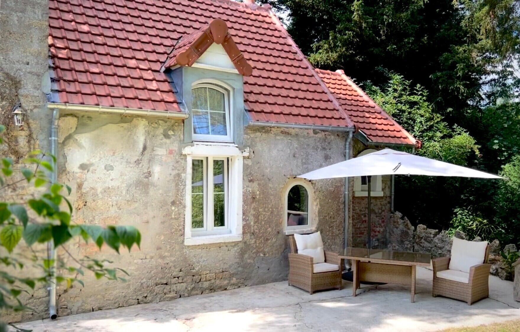 Ein rustikales Steinhaus mit rotem Ziegeldach, einem kleinen Fenster und einer Terrasse mit Korbmöbeln und einem weißen Sonnenschirm.