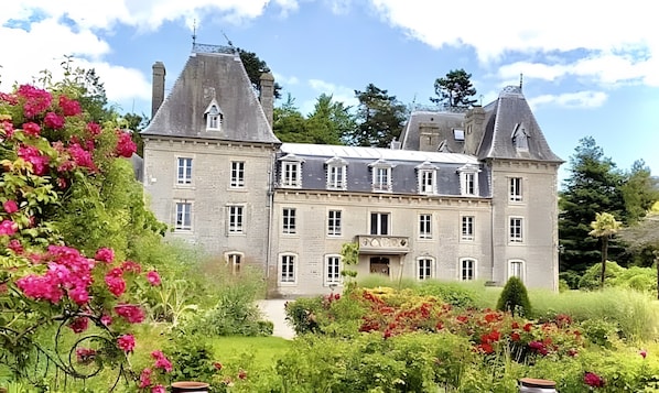 A large, historic building with multiple towers and a central entrance, surrounded by lush gardens and flowering plants.