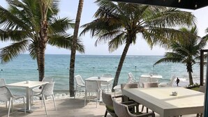 A beachside patio with tables and chairs, palm trees, and a view of the ocean. - Award-Winning Beachfront Resort Penthouse | Pools, Dining & Kite Beach (Cabarete)