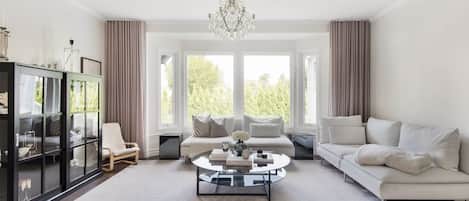 A spacious living room with a large window, a chandelier, a black cabinet, and a white sofa.