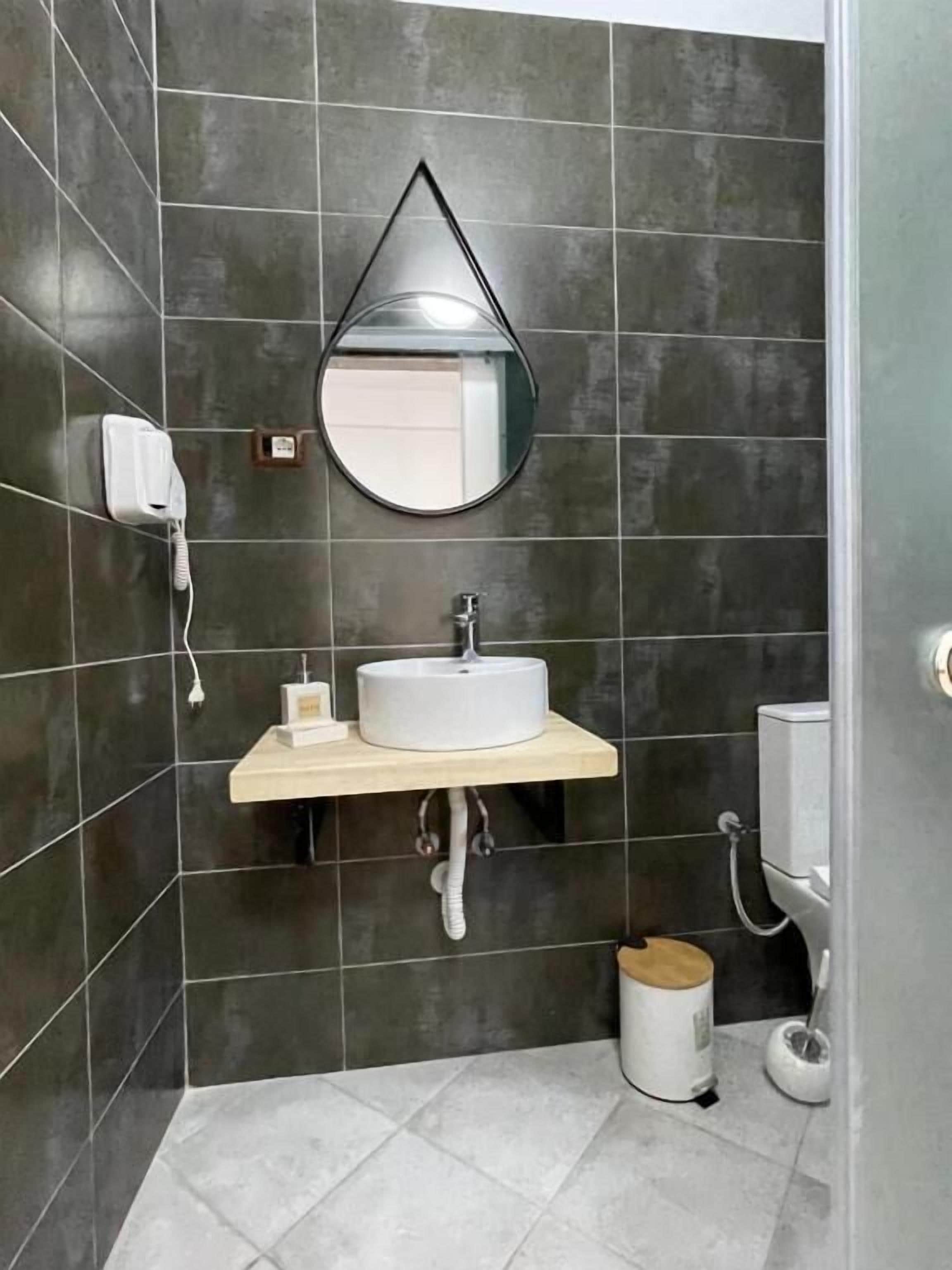 Un baño con azulejos oscuros, un lavamanos blanco, un espejo, un cabezal de ducha de mano y un inodoro.