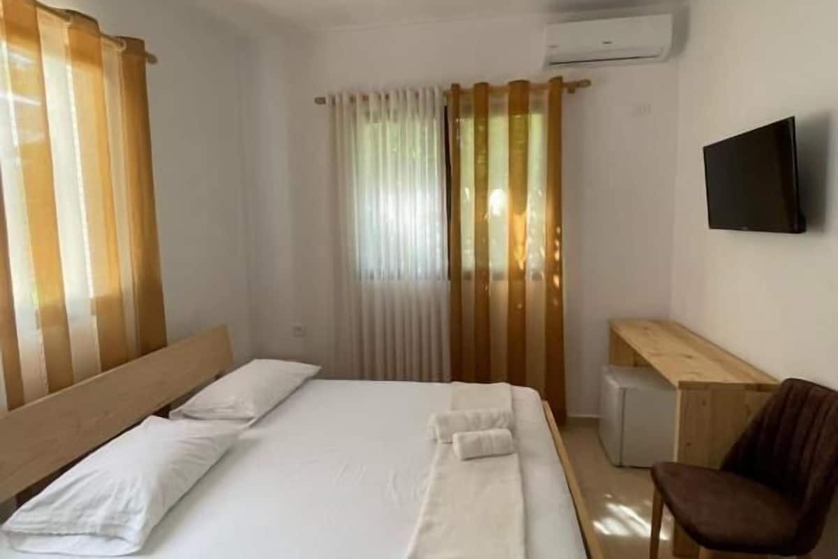 Quarto com cama de madeira, roupa de cama branca, escrivaninha, cadeira e TV fixada na parede.