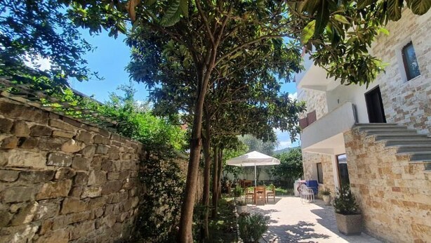Un patio con paredes de piedra, una mesa y sillas bajo un paraguas blanco, una escalera que conduce a un segundo piso y vistas de una colina al fondo.