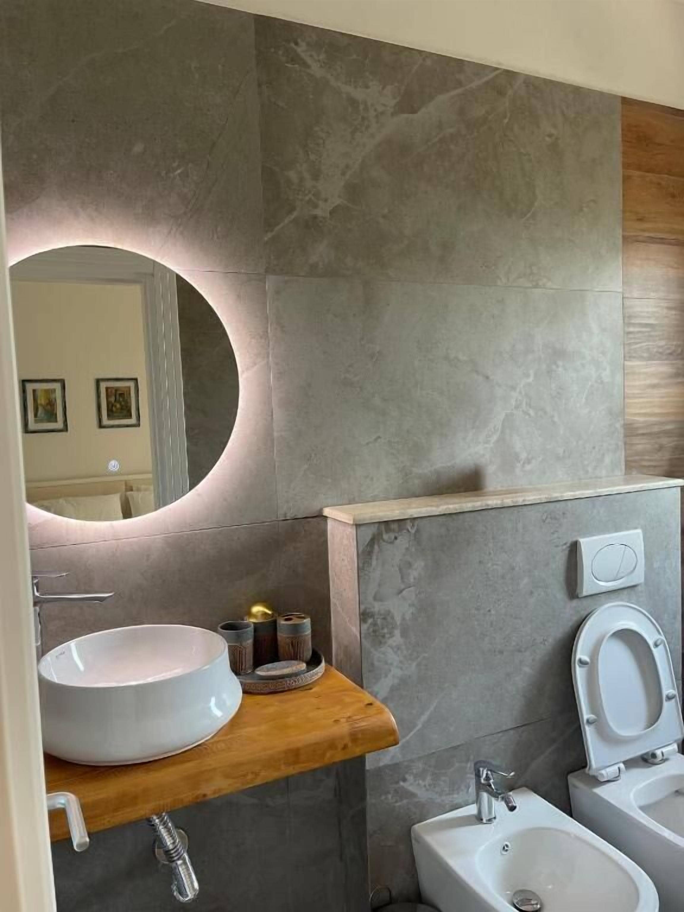 A modern bathroom with a round mirror, a wooden vanity, a bidet, and a sink.
