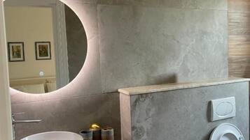 A modern bathroom with a round mirror, a wooden vanity, a bidet, and a sink.