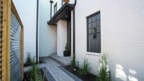 Un bâtiment moderne aux murs en briques blanches, doté d’une porte en bois et d’un chemin pavé agrémenté de plantes.