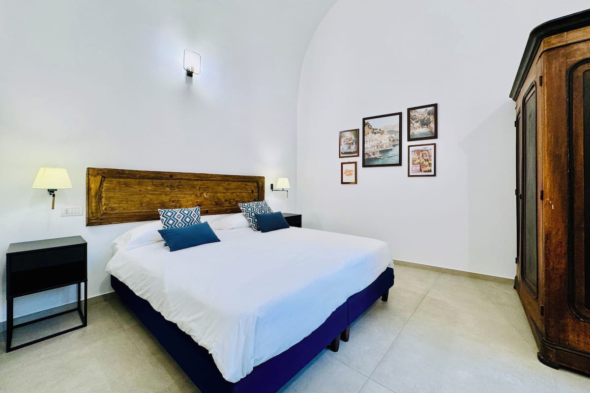 Quarto com cabeceira de madeira, cama com roupa de cama branca e azul, duas luminárias de cabeceira, mesa de cabeceira e guarda-roupa.