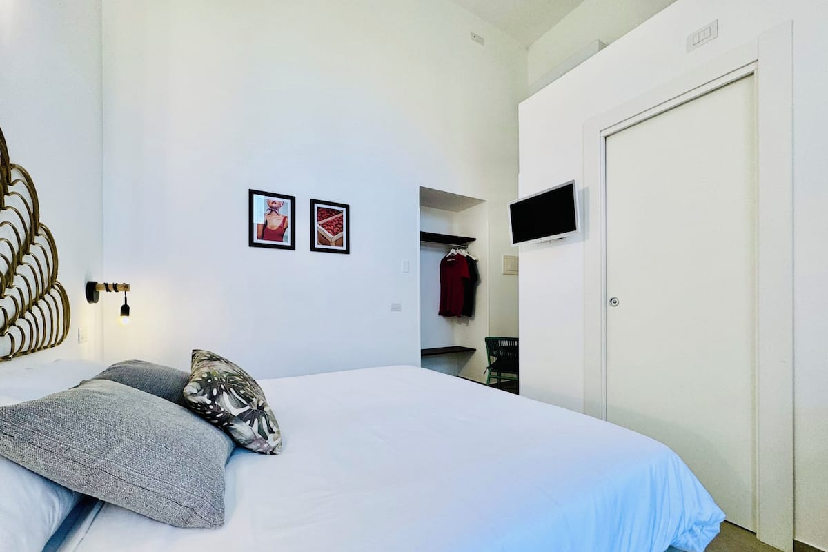 Quarto com cama, televisão na parede, porta e cabideiro com roupas.
