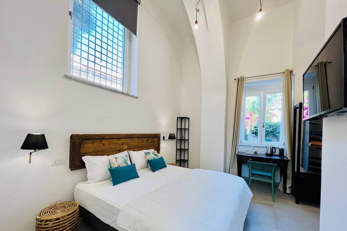 Quarto com cama, luminária, janela, escrivaninha e televisão.