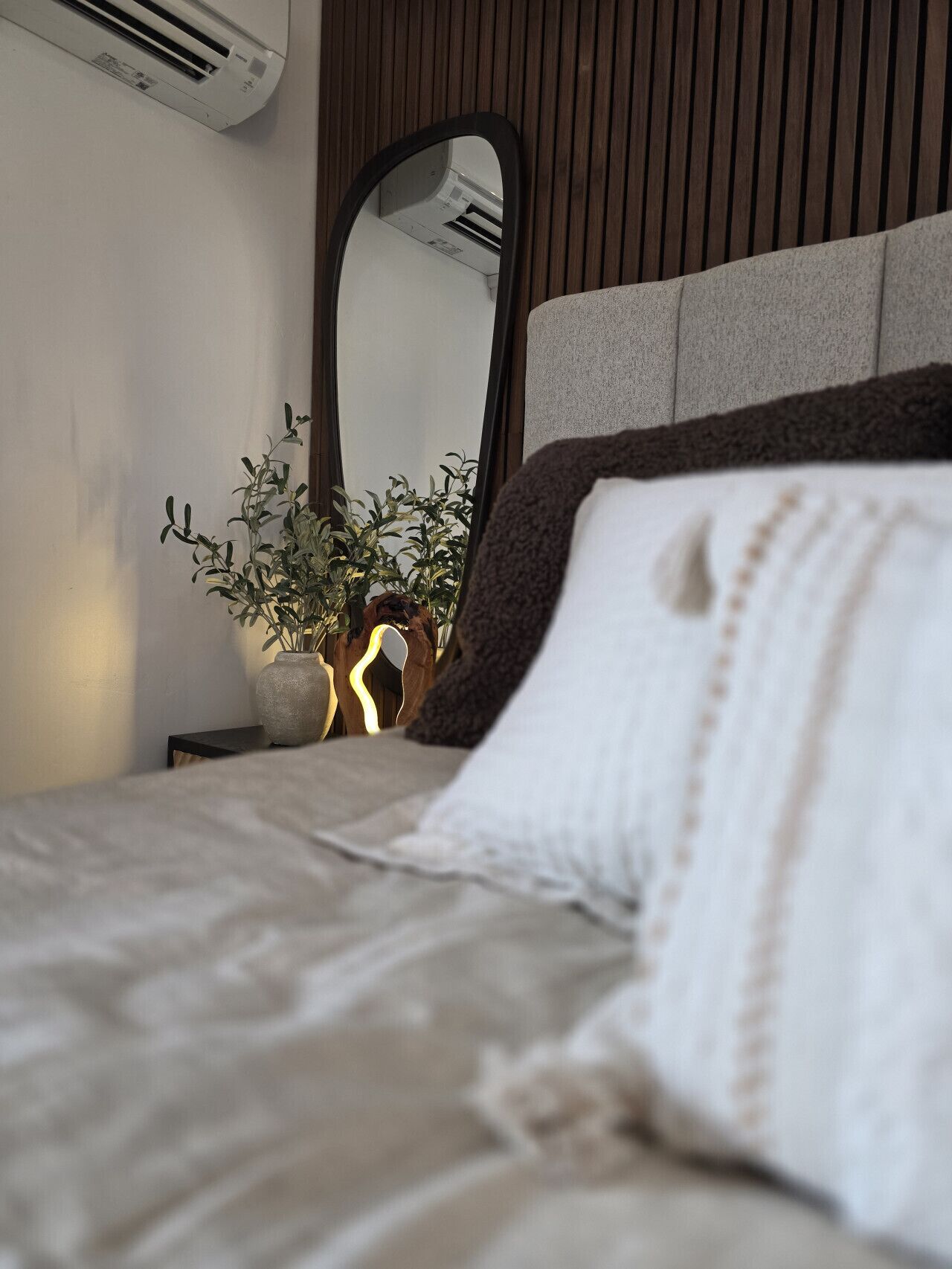 A bed with a white pillow, a potted plant, and a mirror on a wall.