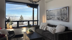 A living room with a large window, a chandelier, a sofa, a coffee table, and a painting on the wall. - Luxury home with hot tub (Victor)