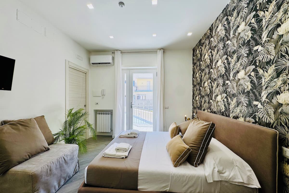 A bedroom with a bed, two sofas, a radiator, an air conditioner, a TV, and a view of the outside.