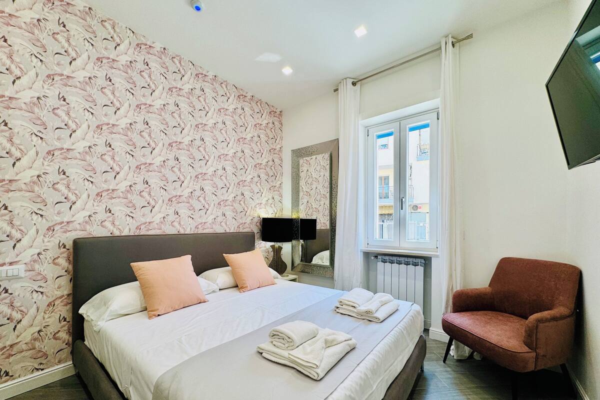 A hotel room with a bed, a chair, a window, and a wall with a floral pattern.