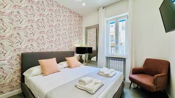 A hotel room with a bed, a chair, a window, and a wall with a floral pattern.