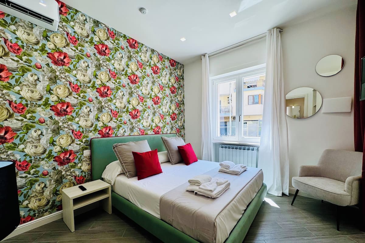 A bedroom with a floral wallpaper, a green headboard, a bed with white and red bedding, a bedside table, a chair, and a window with curtains.