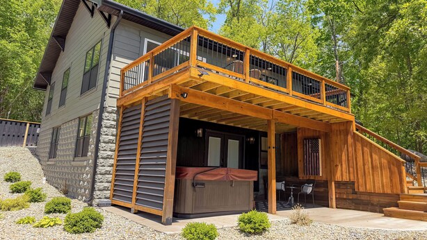 A two-story house with a wooden deck, a hot tub, and a stone wall.