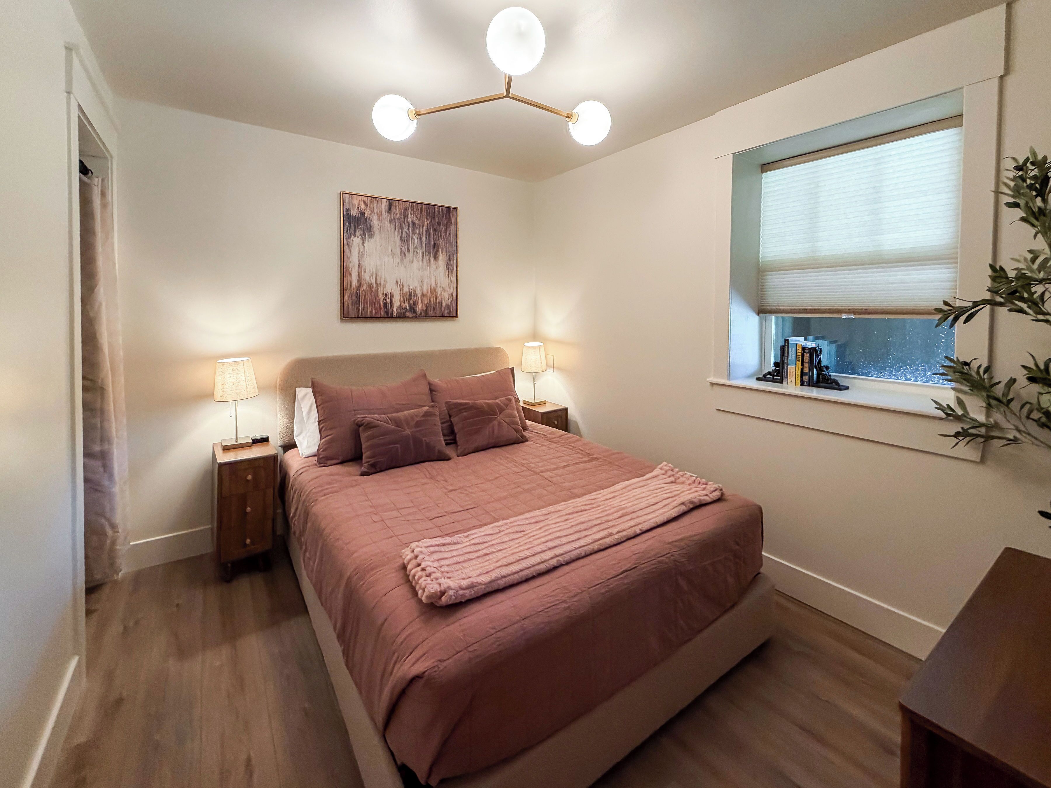 A bedroom with a bed, bedside lamps, a ceiling light fixture, a window with blinds, and a painting on the wall.