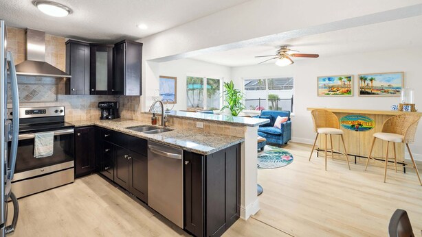 A modern kitchen with dark cabinetry, a stainless steel range, and a built-in dishwasher. There is a dining area with a bar counter and two bar stools. The room has a ceiling fan and large windows. - St. Pete Pool Oasis - Steps to the Beach! (Pinellas Park)