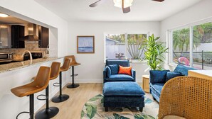 A modern kitchen with dark cabinetry, a countertop, and a ceiling fan. There are three bar stools and a blue sofa with a cushion. A large window provides a view of palm trees and a swimming pool. - St. Pete Pool Oasis - Steps to the Beach! (Pinellas Park)