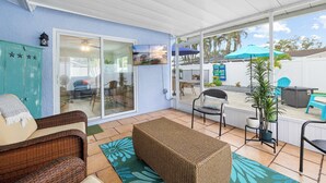 A covered patio with wicker furniture, a teal cabinet, and a sliding glass door leading to an outdoor area with a table, chairs, and umbrellas. - St. Pete Pool Oasis - Steps to the Beach! (Pinellas Park)