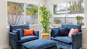 A corner room with two blue armchairs, a small ottoman, a potted plant, and large windows with a view of palm trees and a fence. - St. Pete Pool Oasis - Steps to the Beach! (Pinellas Park)