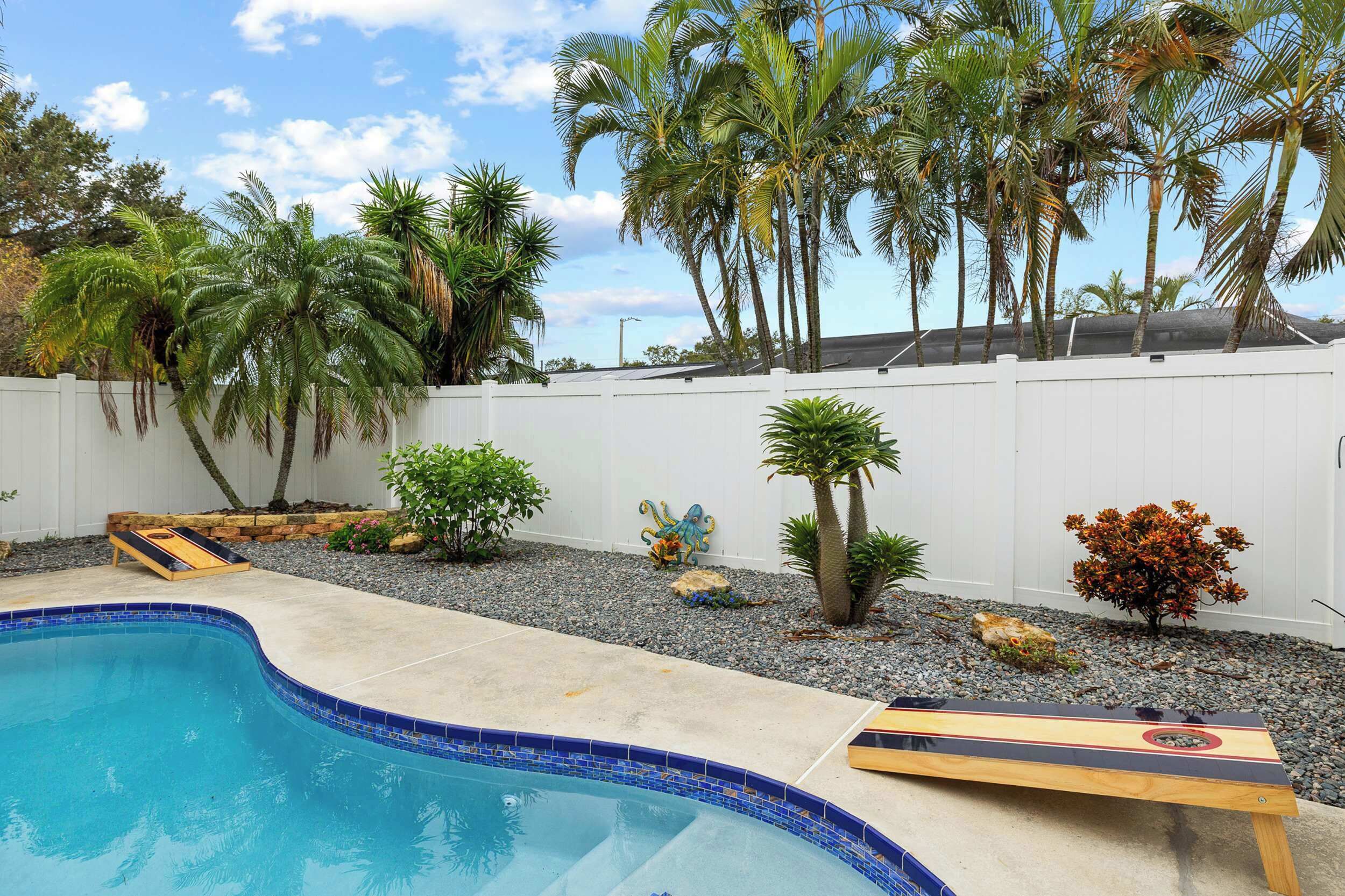 Una piscina con borde de azulejos, rodeada de palmeras y una valla blanca.