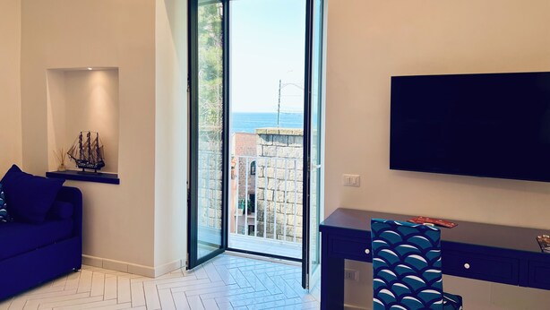 A modern living room with a sofa, a desk, a chair, a TV, and a large window with a view of the sea. - YourHome - Scirocco Suite (Sorrento)