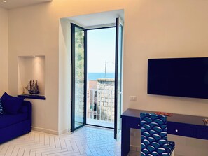 A modern living room with a sofa, a desk, a chair, a TV, and a large window with a view of the sea. - YourHome - Scirocco Suite (Sorrento)