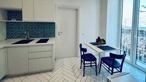 A compact kitchen with a sink, cabinets, and a tiled backsplash. There is a dining area with a table and chairs, and a view through a large window. - YourHome - Scirocco Suite (Sorrento)
