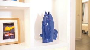 Eine blaue Skulptur auf einem weißen Regal, ein gerahmtes Bild von einem Fisch und ein gelbes Werkzeug.