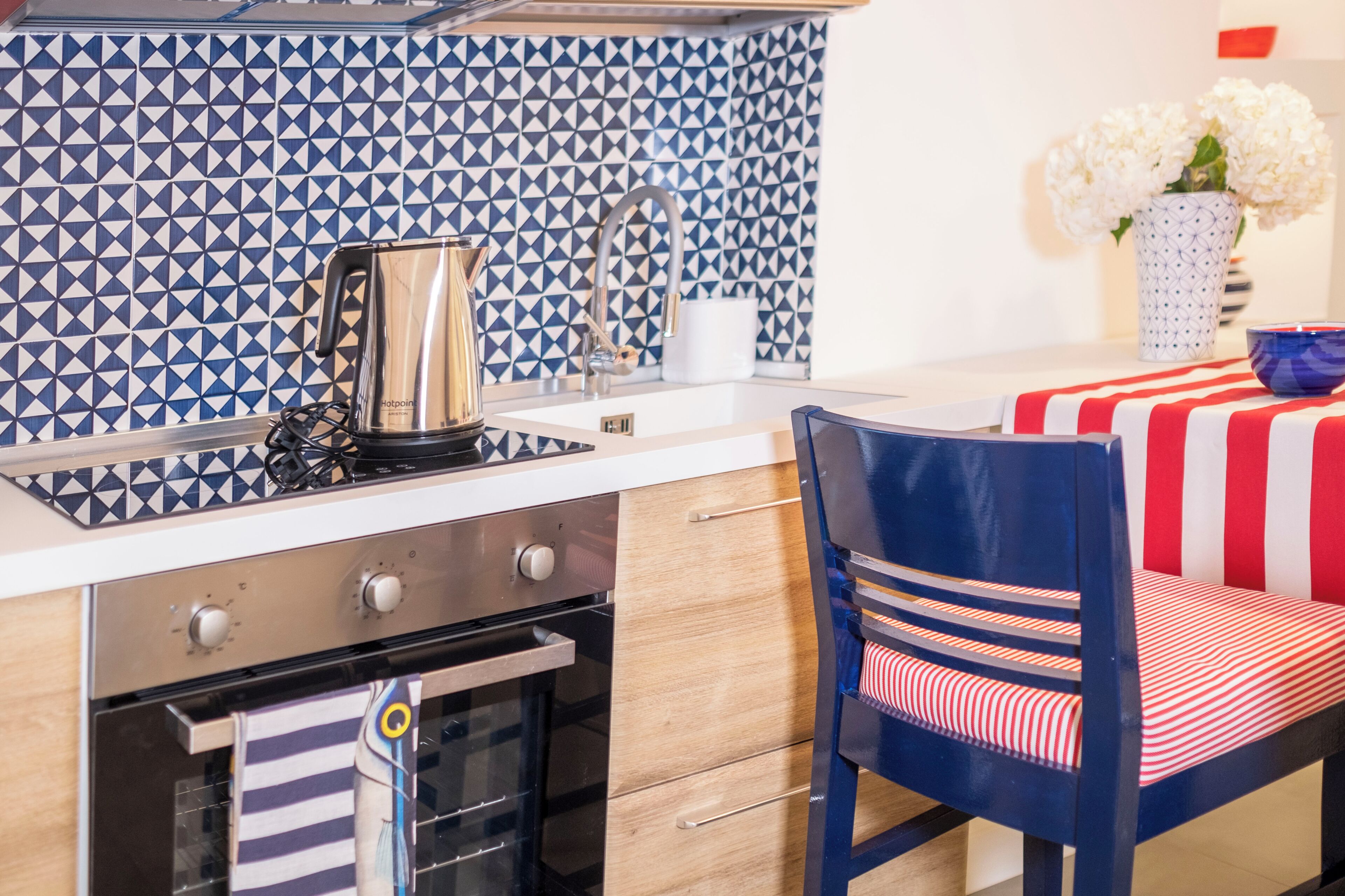 A kitchen with a patterned backsplash, a coffee maker, a sink, and a dining area with a striped tablecloth and a chair.