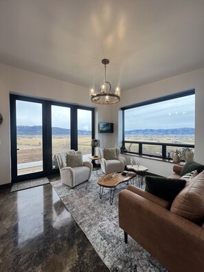 A spacious living room with a large window, a chandelier, and a flat-screen TV. - Luxury home with hot tub (Victor)