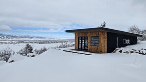 Moderni puinen mökki, jossa on suuri lasiovi, sijaitsee lumisessa maisemassa kirkkaan taivaan alla.