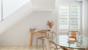 Un coin repas comprenant une table en verre, des chaises, un bureau en bois et une fenêtre avec des stores.
