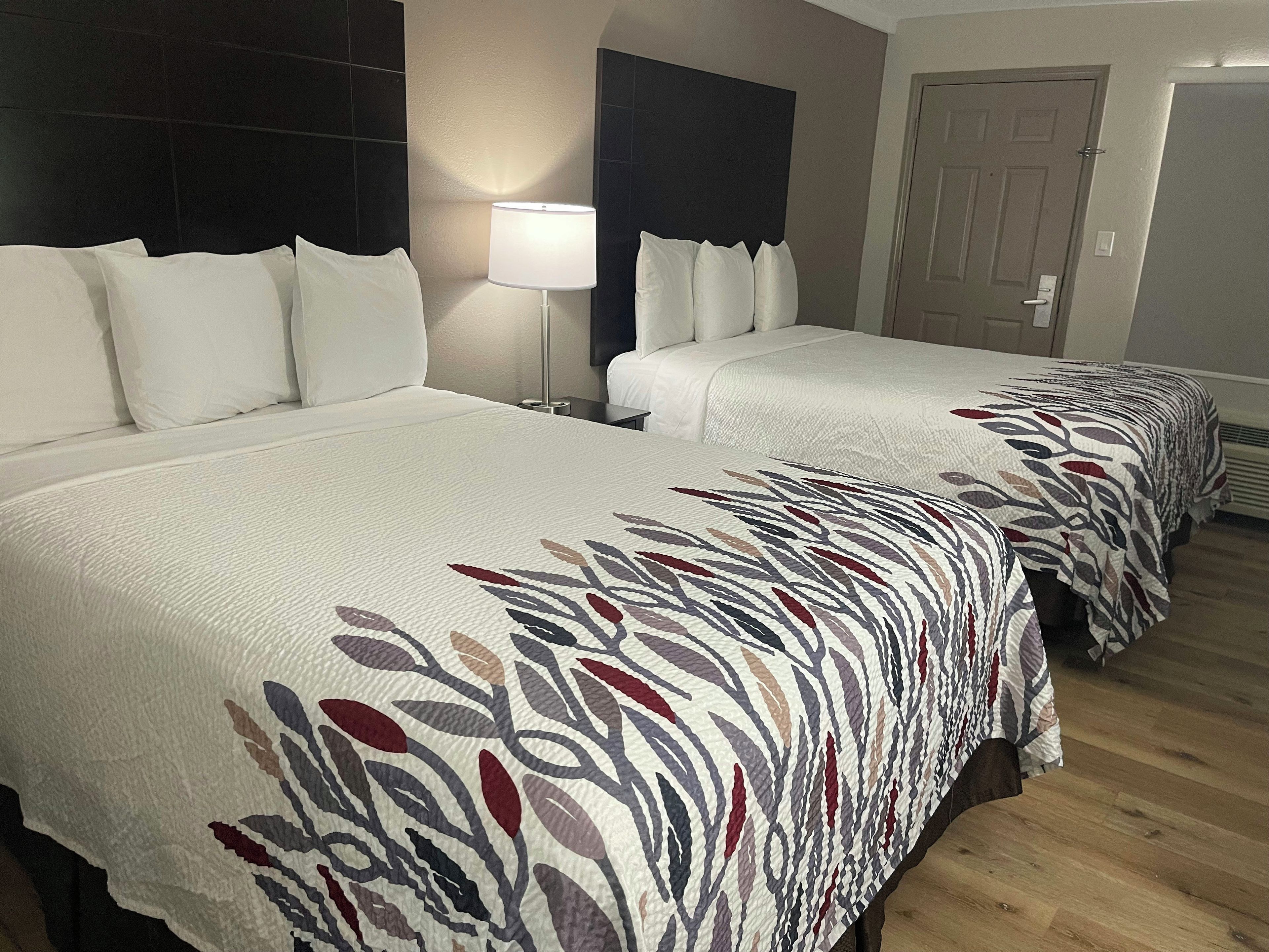Quarto de hotel com duas camas, uma mesa de cabeceira, uma luminária e uma porta.