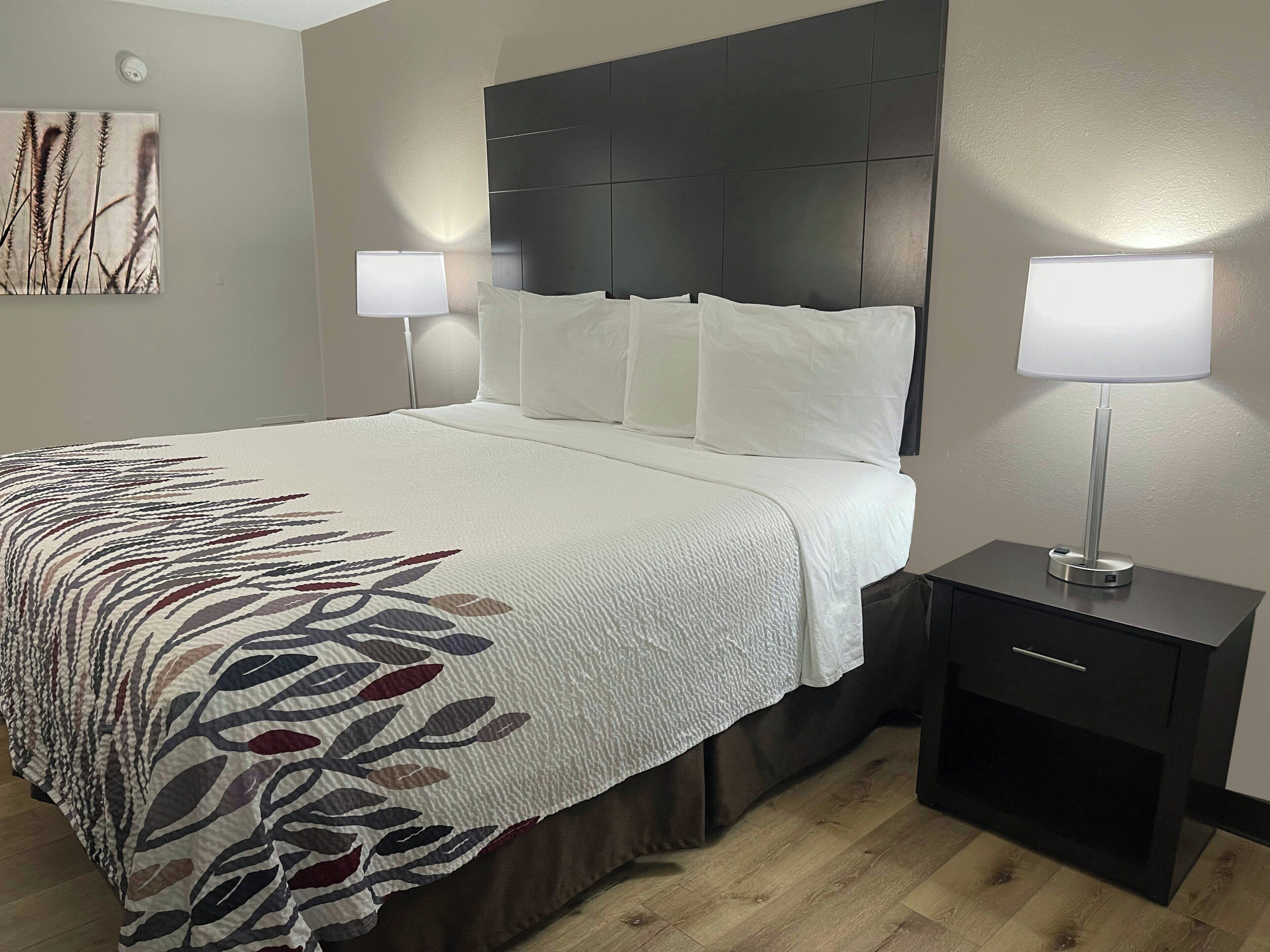 Una cama bien hecha con ropa blanca y un plaid estampado, un cabecero oscuro, dos lámparas de noche y una mesita de noche con una lámpara.