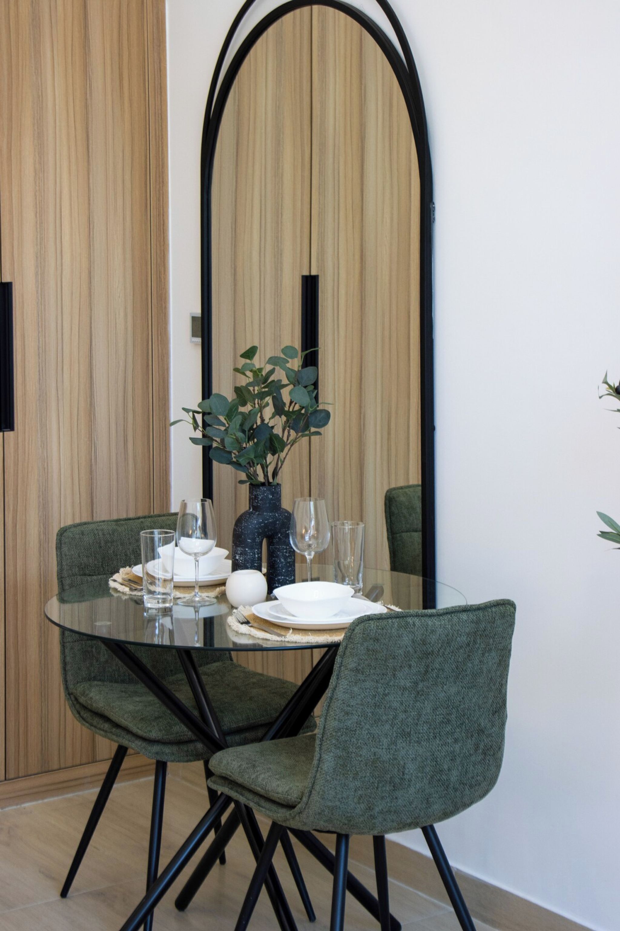 A dining area with a round glass table, two upholstered chairs, a vase with green leaves, and a mirror on the wall.