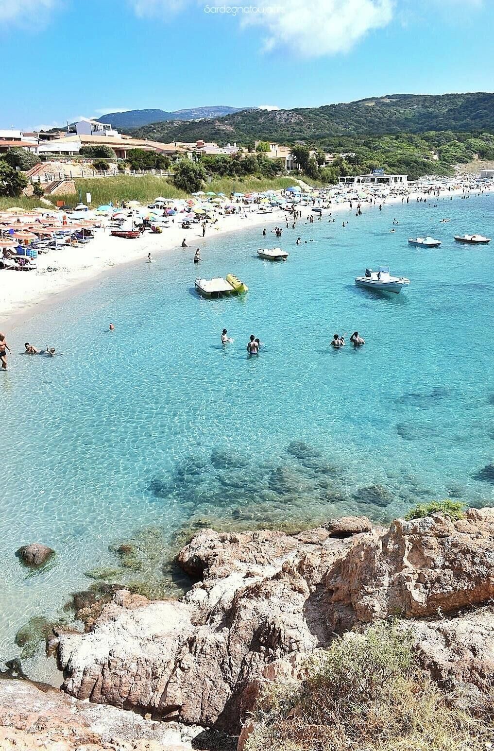 Una playa costera con agua cristalina, embarcaciones y un primer plano rocoso.