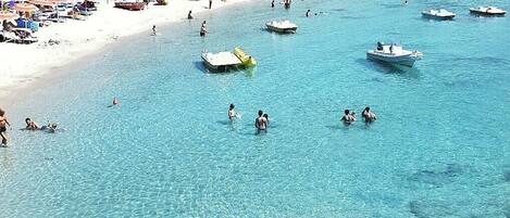 Una playa costera con agua cristalina, embarcaciones y un primer plano rocoso.