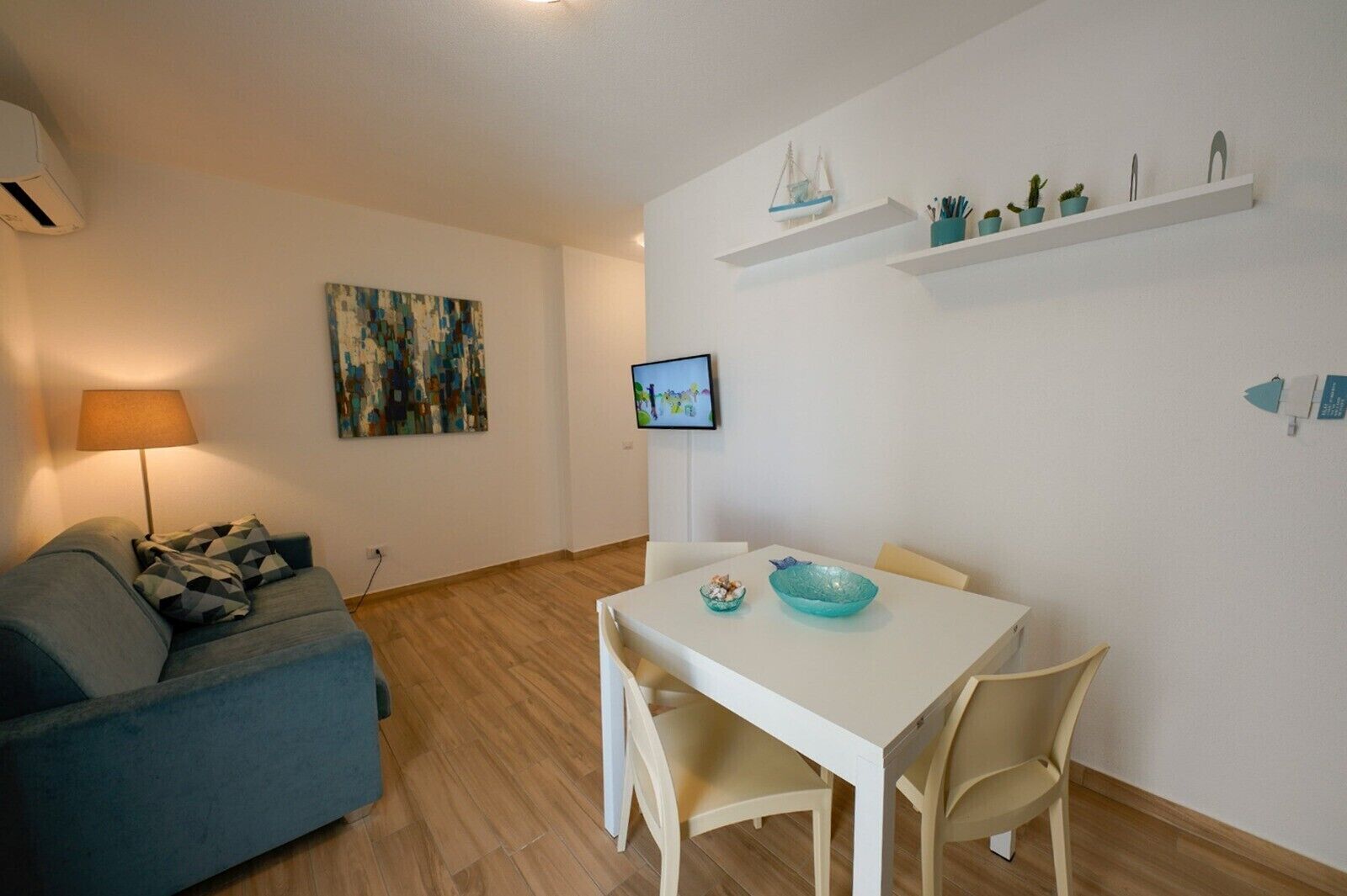 Ett vardagsrum med en soffa, ett bord med stolar, en tv, en tavla och en hylla med dekorativa föremål.