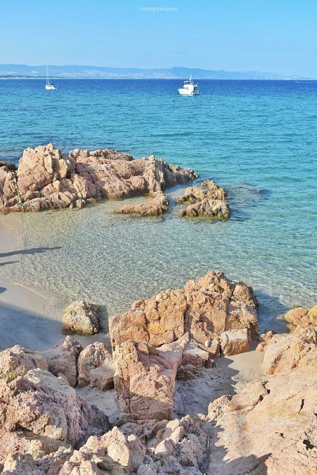 A rocky shoreline with clear water, boats in the distance, and a clear sky.