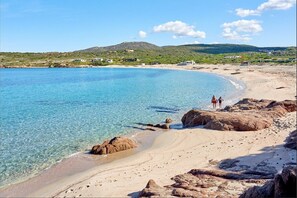 Ein Strand mit klarem Wasser, felsigem Ufer und Bergen in der Ferne.