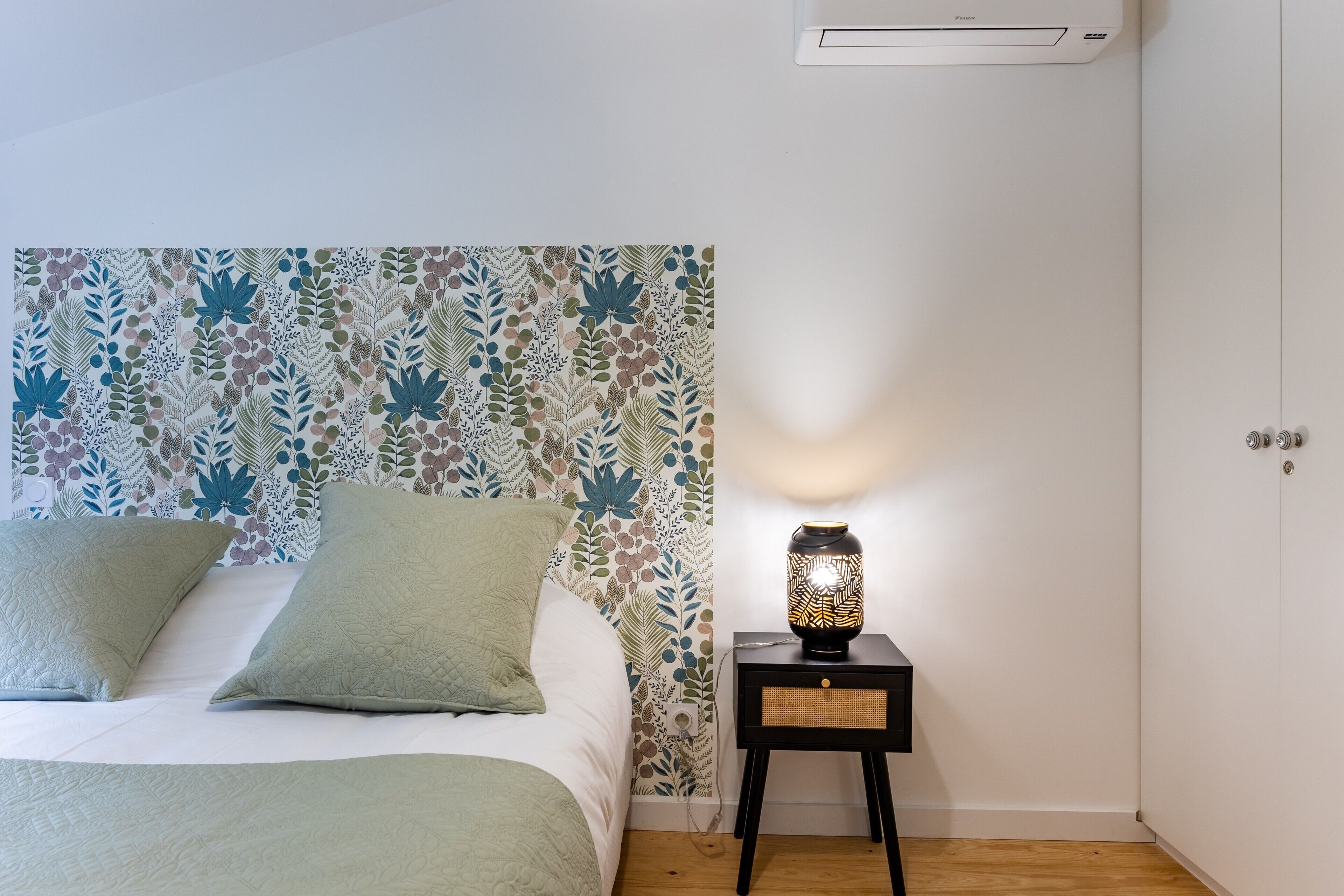 Een slaapkamer met behang met patroon, een bed met groene beddengoed, een nachtkastje met een lamp, en een witte airconditioning.