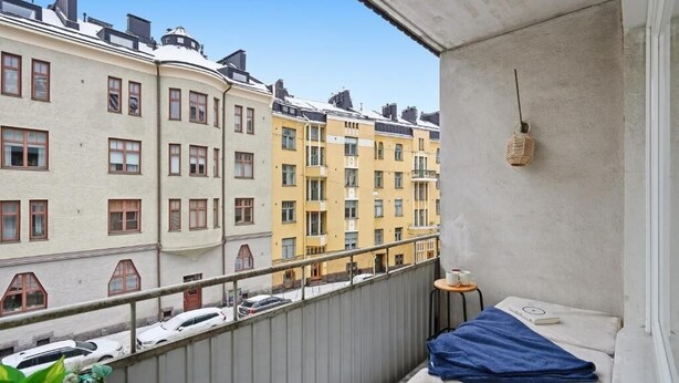 Parveke, jolta avautuu näkymä korkeisiin kerrostaloihin, pieni pöytä, jossa on kuppi, ja riippuvalaisin.